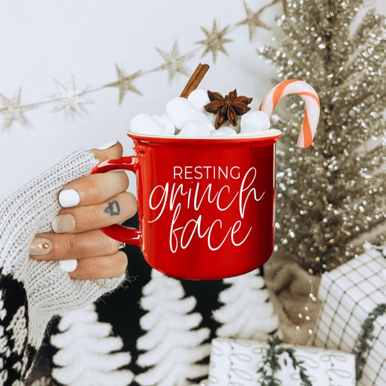 A whimsical Grinch Face Mug in red ceramic, featuring the Grinch's face and white lettering, perfect for holiday beverages.