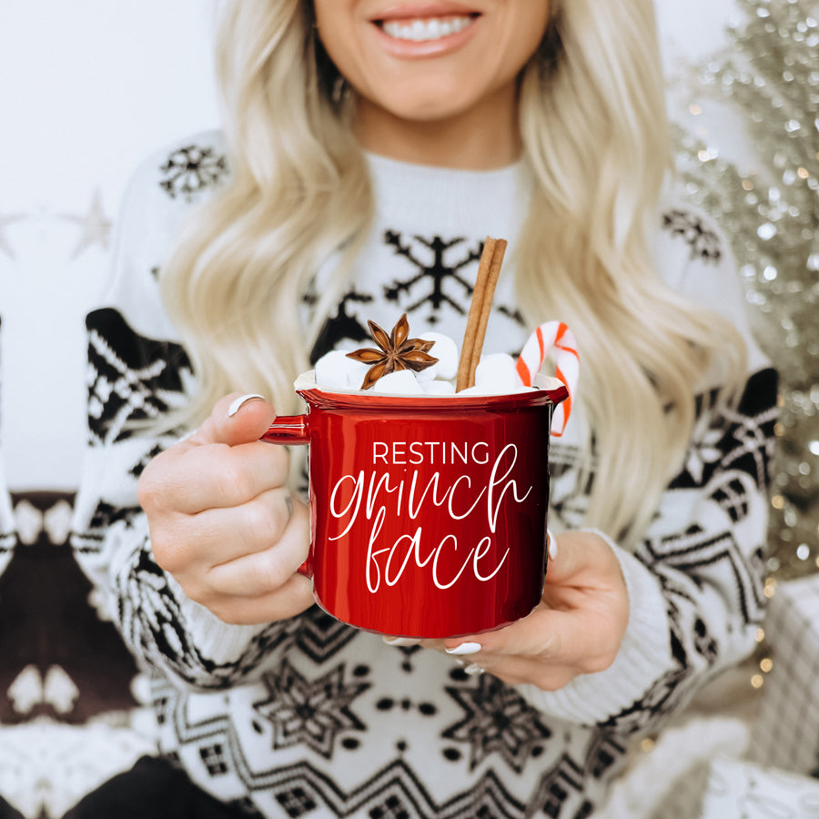 A whimsical Grinch Face Mug in red ceramic, featuring the Grinch's face and white lettering, perfect for holiday beverages.