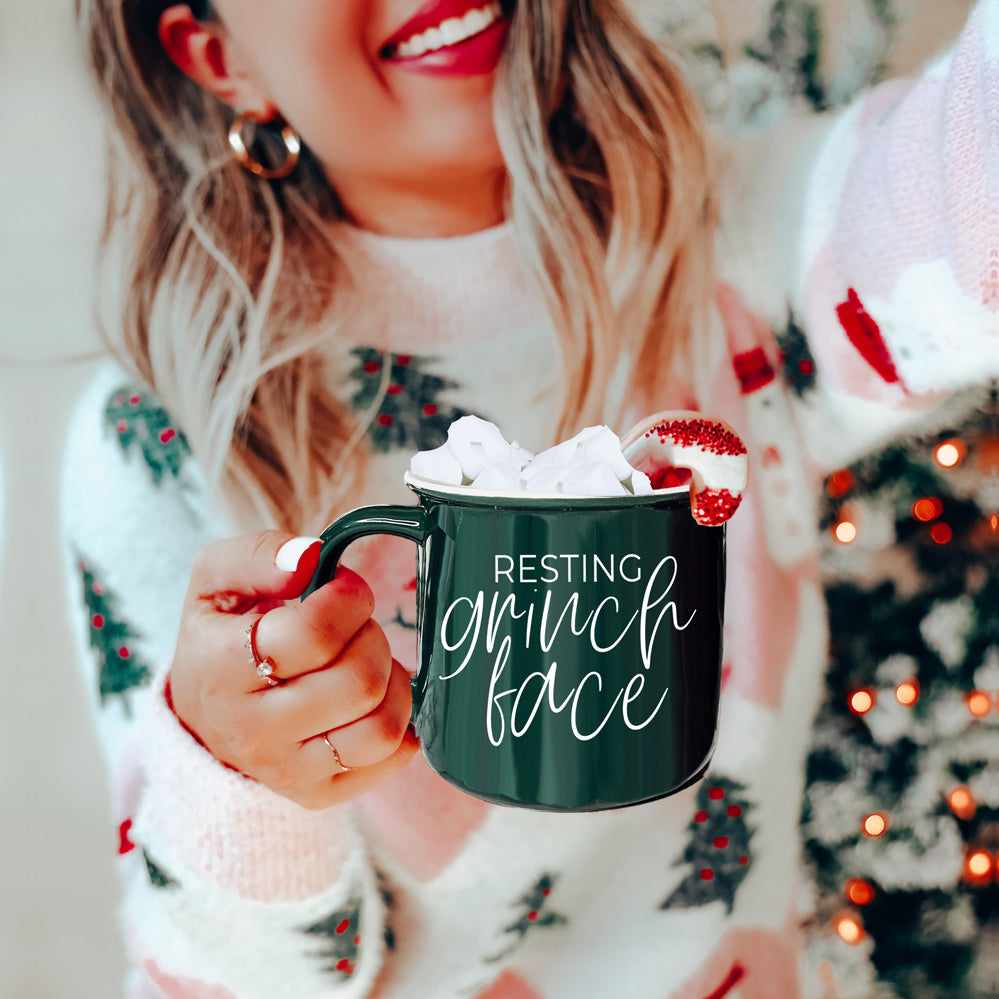 A whimsical Grinch Face Mug in red ceramic, featuring the Grinch's face and white lettering, perfect for holiday beverages.