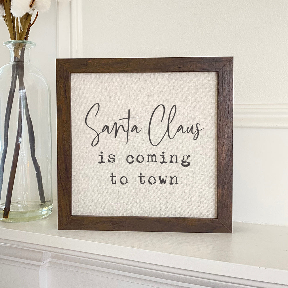 A beautifully framed sign reading 'Santa Claus is Coming to Town' with a rustic wood frame, displayed on a shelf.