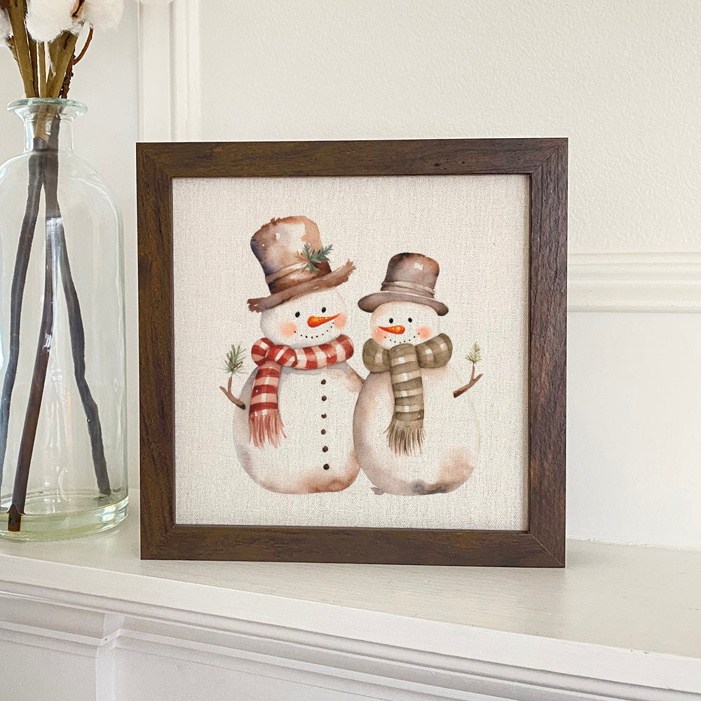 A charming framed sign featuring two snowmen, set in a stylish wood frame, perfect for holiday decor.