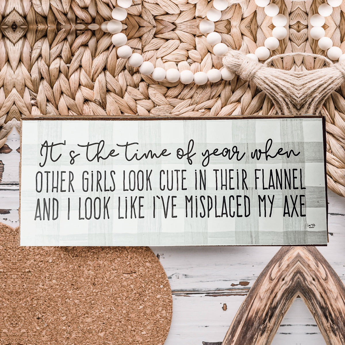 A humorous wooden sign with a flannel design reading 'It's the time of year when other girls look cute in their flannel and I look like I've misplaced my axe.'