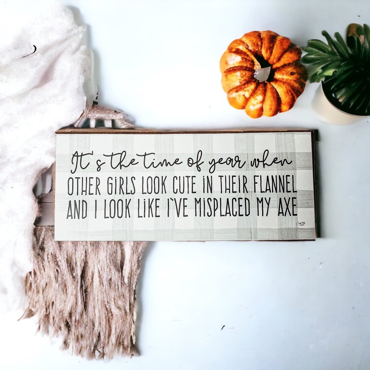 A humorous wooden sign with a flannel design reading 'It's the time of year when other girls look cute in their flannel and I look like I've misplaced my axe.'