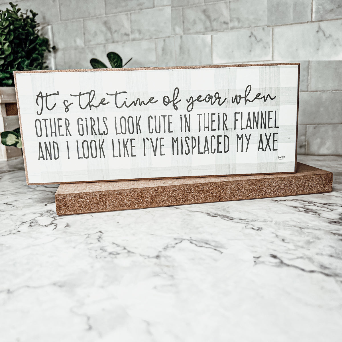 A humorous wooden sign with a flannel design reading 'It's the time of year when other girls look cute in their flannel and I look like I've misplaced my axe.'