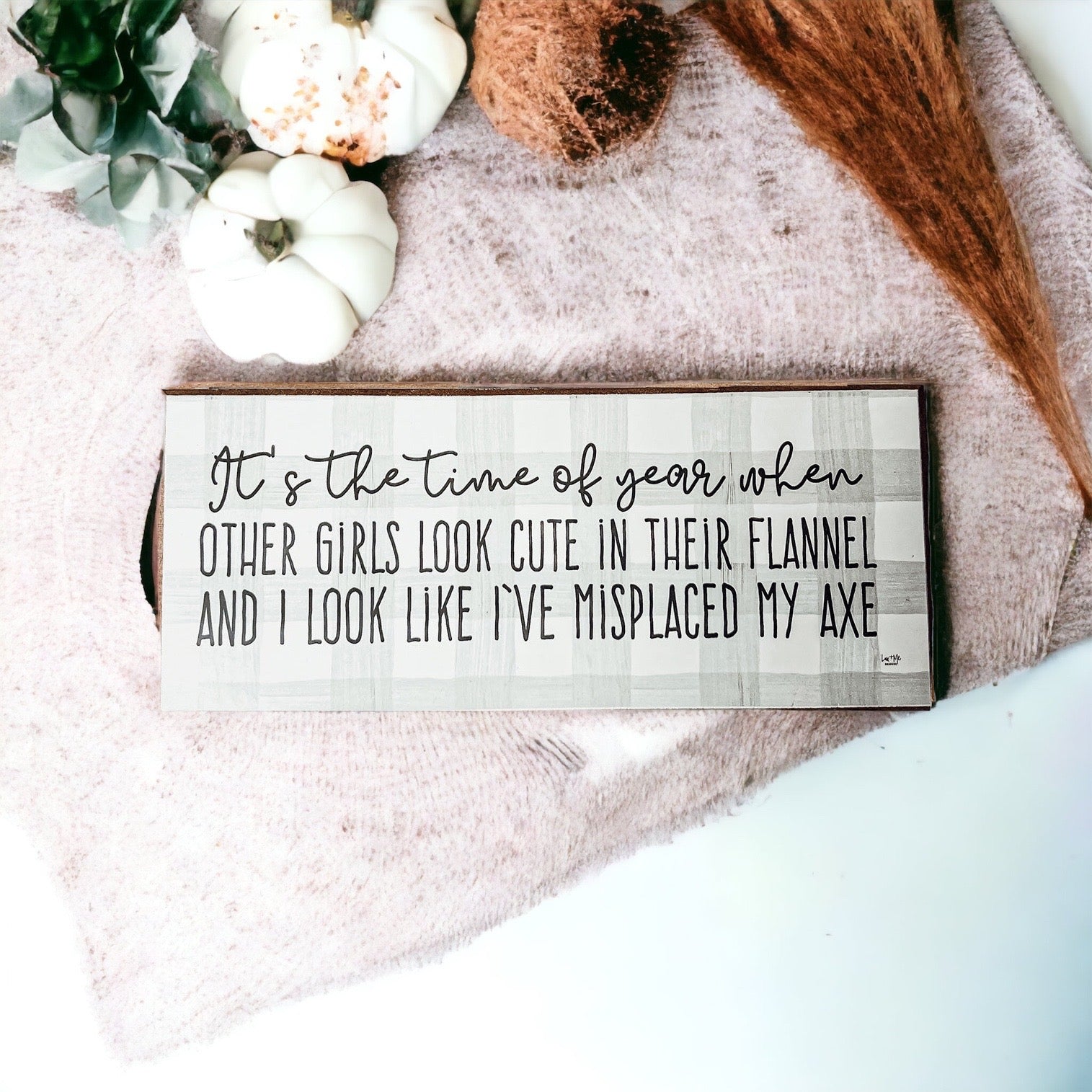 A humorous wooden sign with a flannel design reading 'It's the time of year when other girls look cute in their flannel and I look like I've misplaced my axe.'