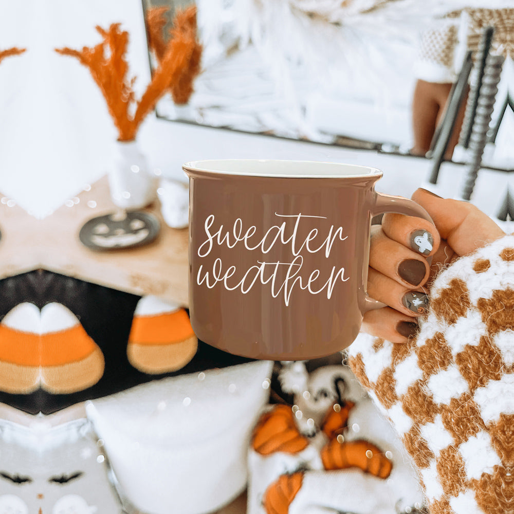 A cozy ceramic campfire style mug in neutral brown with white lettering that says 'Sweater Weather' on both sides.