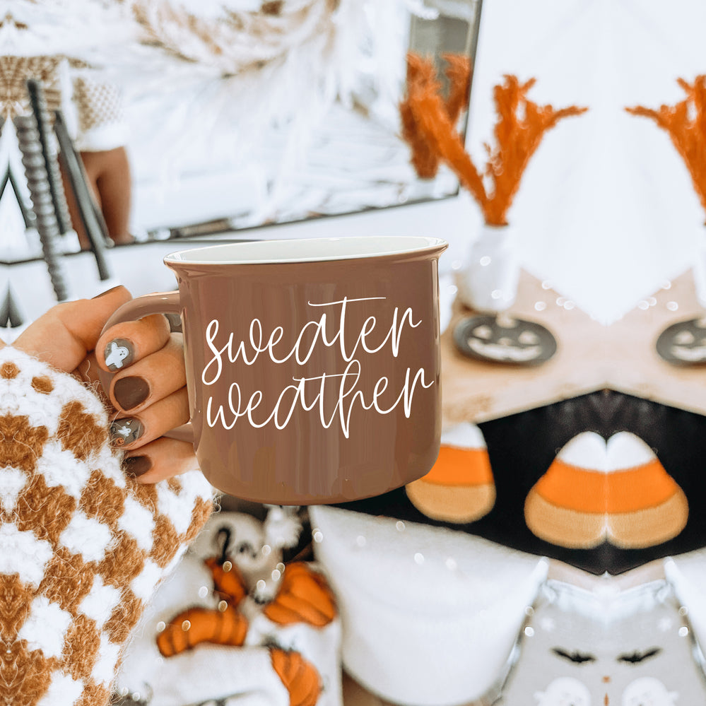 A cozy ceramic campfire style mug in neutral brown with white lettering that says 'Sweater Weather' on both sides.