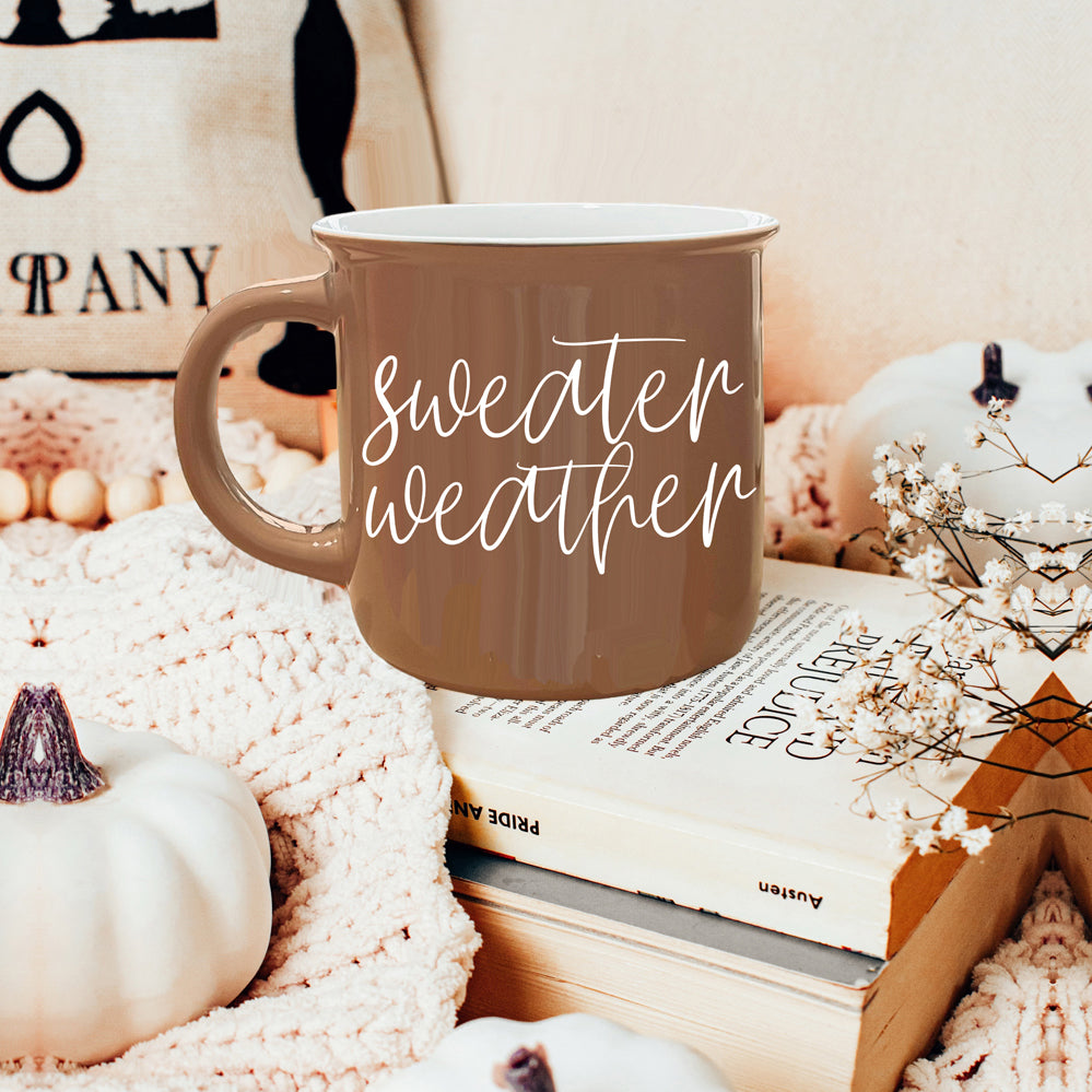 A cozy ceramic campfire style mug in neutral brown with white lettering that says 'Sweater Weather' on both sides.