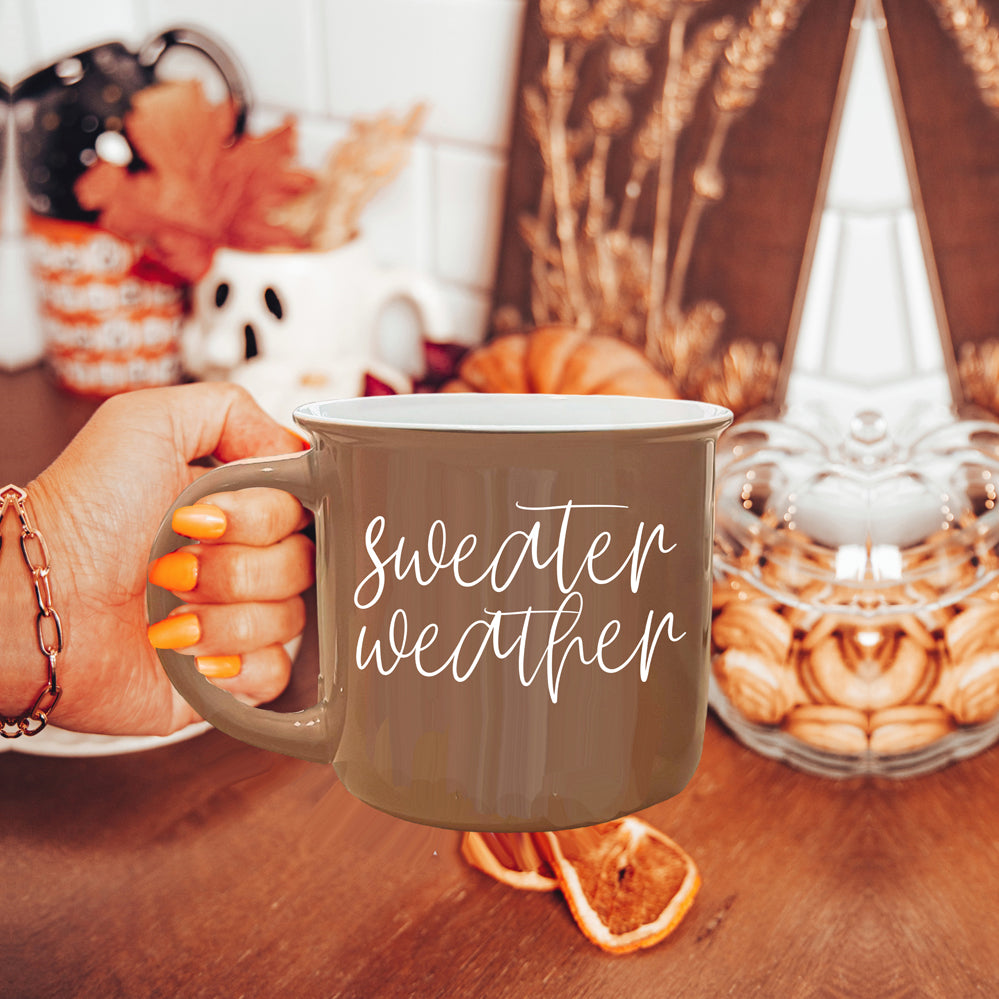 A cozy ceramic campfire style mug in neutral brown with white lettering that says 'Sweater Weather' on both sides.