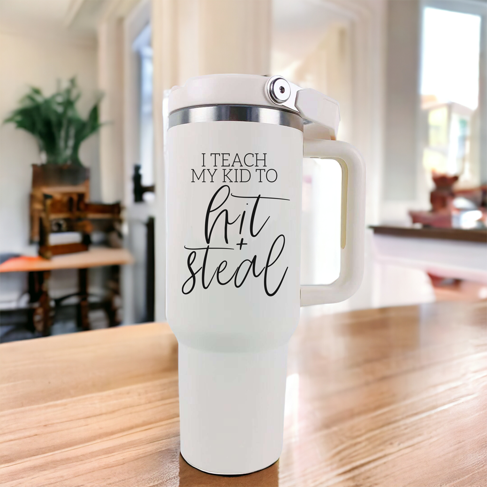 Hit & Steal tumbler with cream exterior and stainless steel interior, featuring a humorous design for baseball moms, double handles, and a no-spill sipping spout.