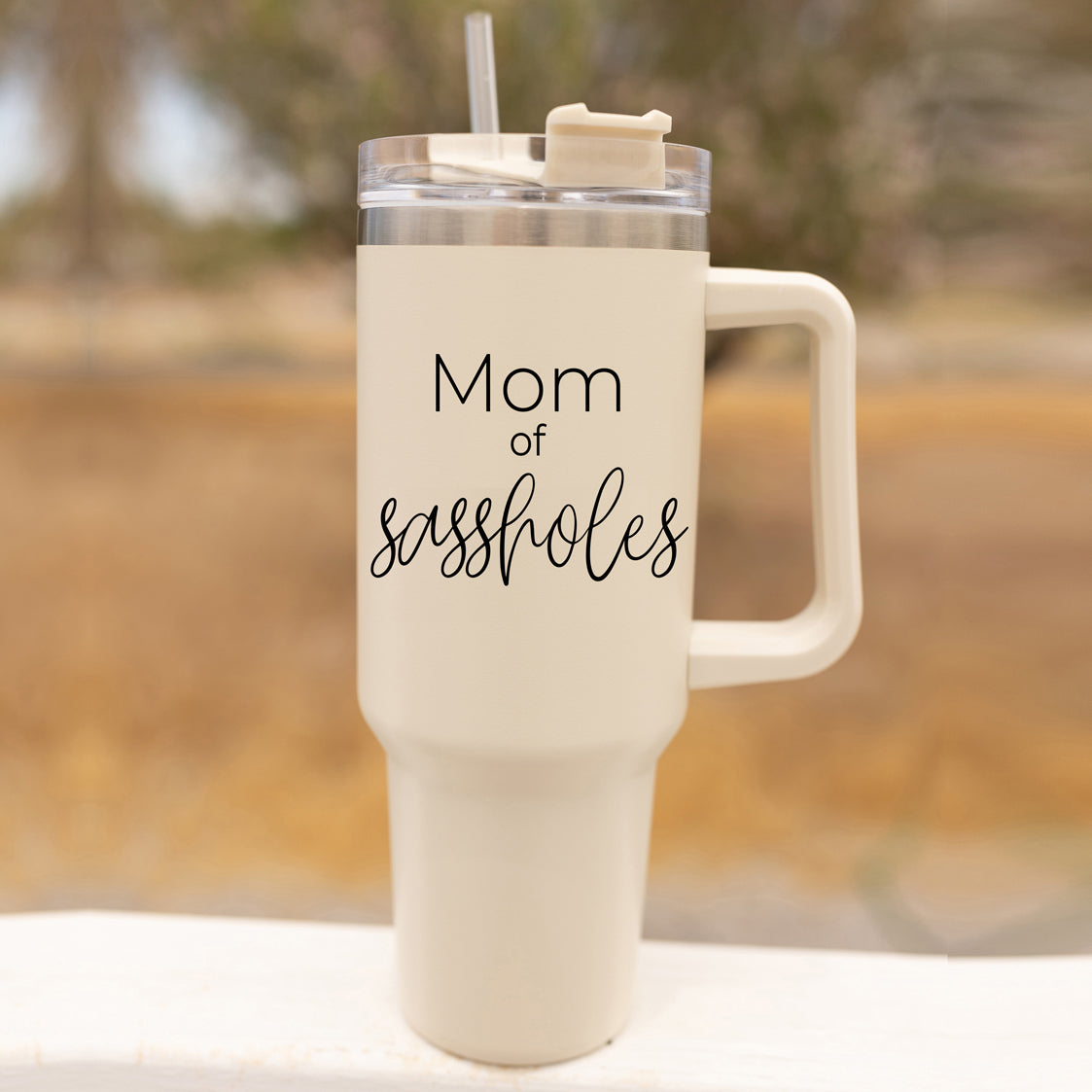 Mom of Sassholes 40oz insulated mug with handle, lid, and straw, featuring a cream exterior and black graphic design.