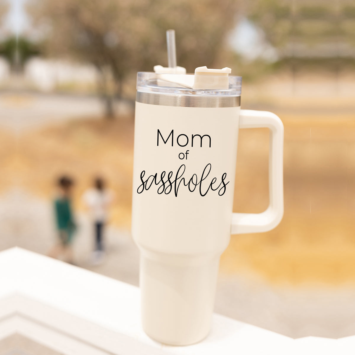 Mom of Sassholes 40oz insulated mug with handle, lid, and straw, featuring a cream exterior and black graphic design.