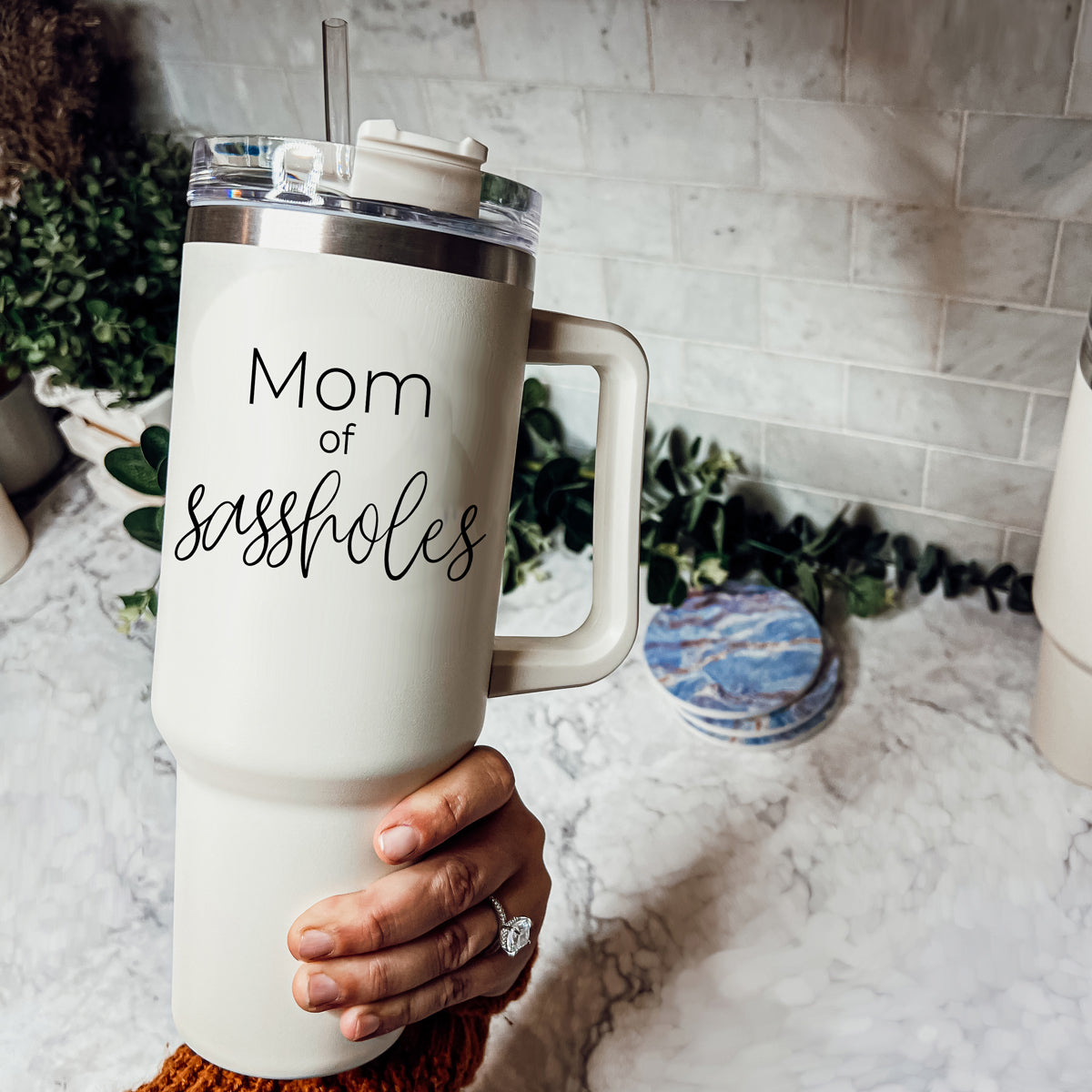 Mom of Sassholes 40oz insulated mug with handle, lid, and straw, featuring a cream exterior and black graphic design.