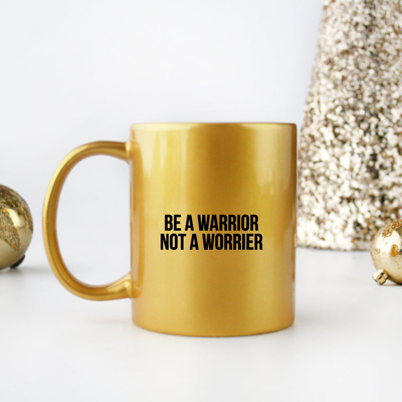 A stylish ceramic mug with gold metallic coating featuring the phrase 'Be A Warrior Not A Worrier' in elegant typography.