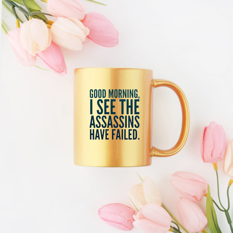 Ceramic mug with gold metallic coating featuring the phrase 'Good Morning I See The Assassins Have Failed' on one or both sides.