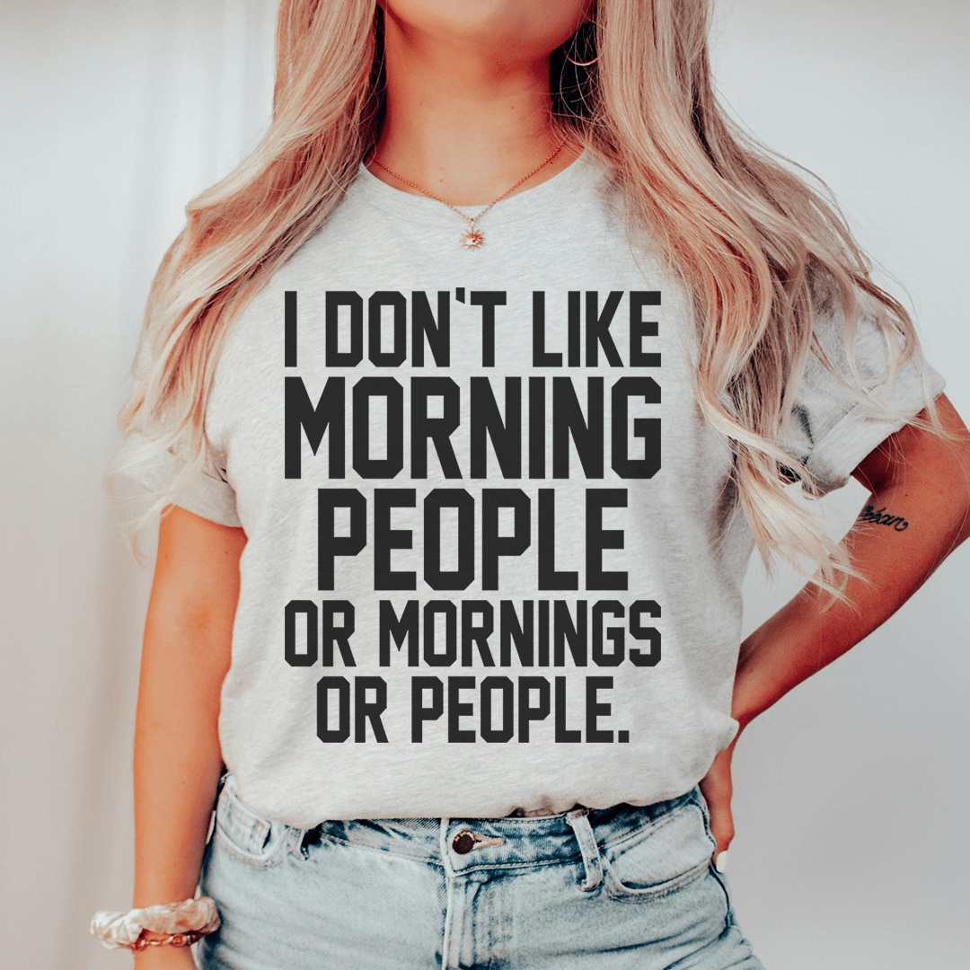 A black t-shirt with the phrase 'I Don't Like Morning People Or Mornings Or People' printed in bold white letters, showcasing a casual and humorous design.