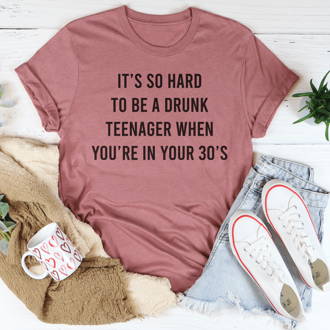 A stylish t-shirt featuring the phrase 'It's So Hard To Be A Drunk Teenager When You're In Yours 30's', made from soft cotton with double stitching.