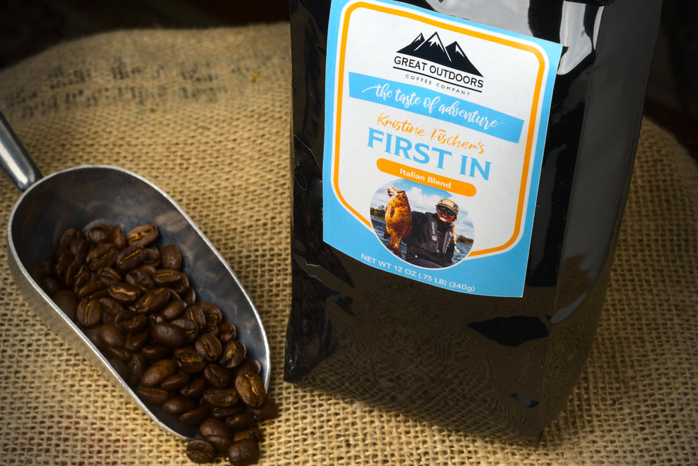 A steaming cup of First In coffee by Kristine Fischer, showcasing its rich color and inviting aroma, set against a serene outdoor backdrop.