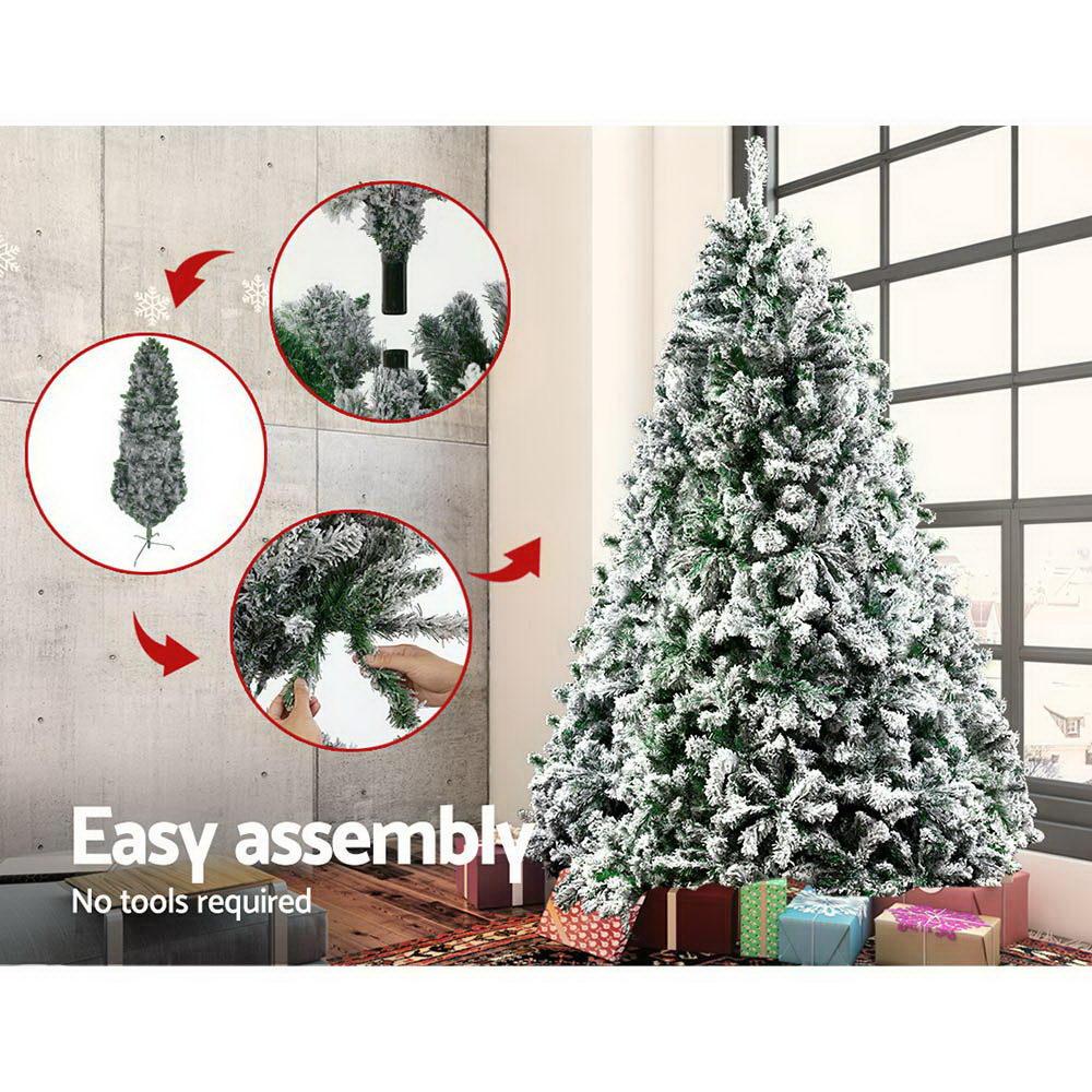 Jingle Jollys 2.1M Christmas Tree with snow-sprayed tips, standing tall in a festive living room setting, adorned with colorful ornaments.