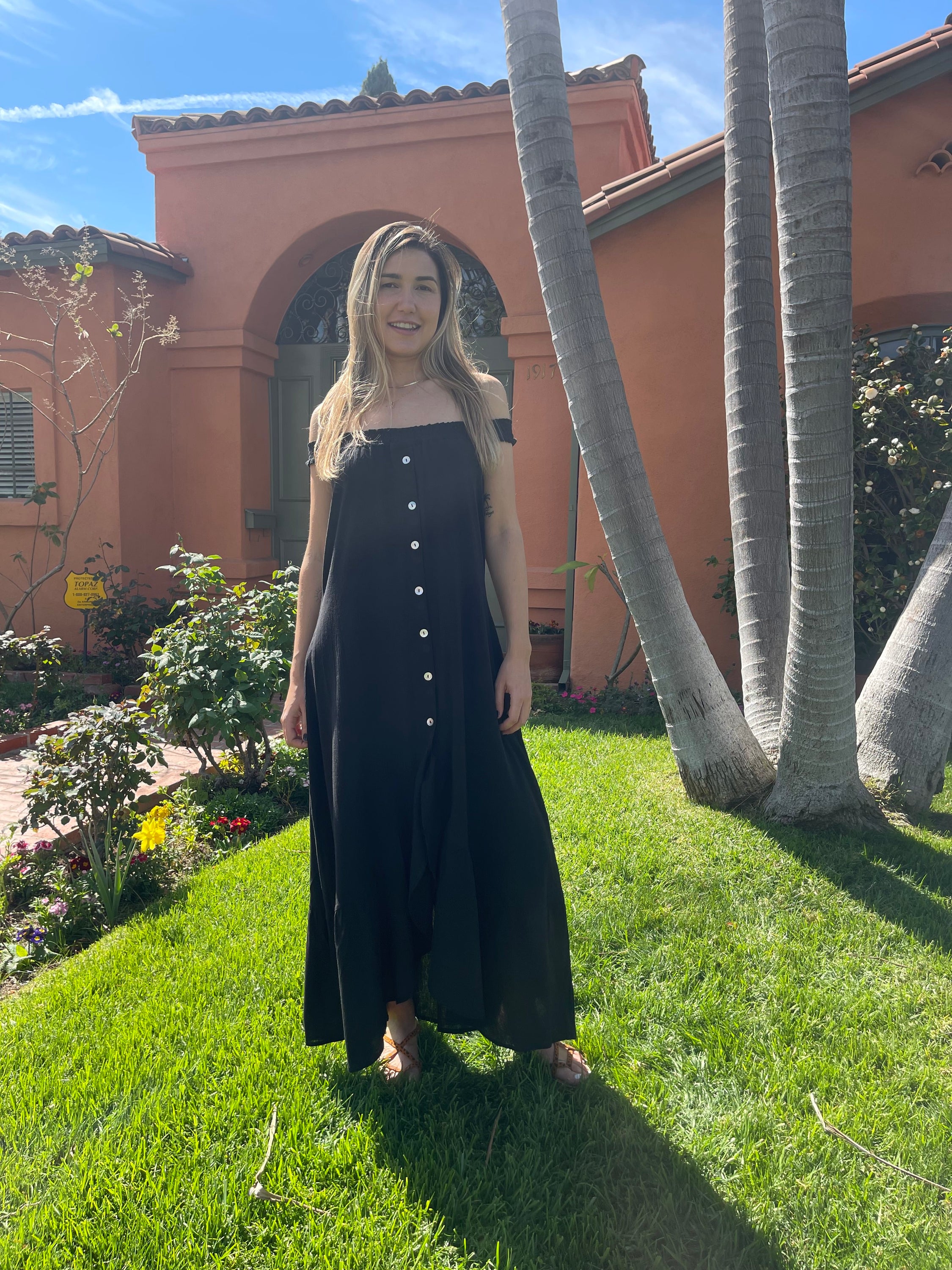 A stylish black MALIBU MAXI DRESS featuring a scooped neckline, shirred detailing, and mother-of-pearl buttons, perfect for sunny occasions.