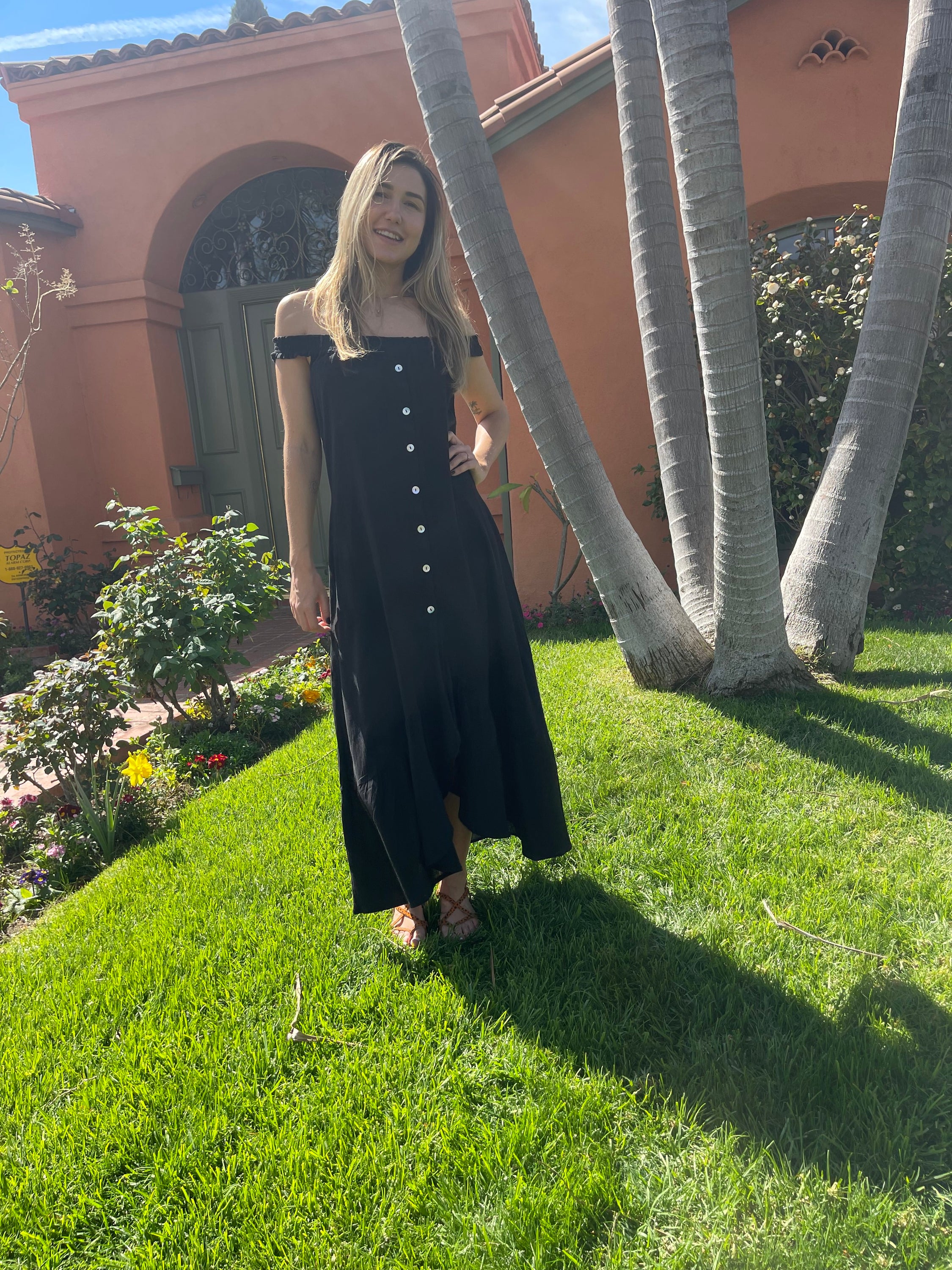 A stylish black MALIBU MAXI DRESS featuring a scooped neckline, shirred detailing, and mother-of-pearl buttons, perfect for sunny occasions.