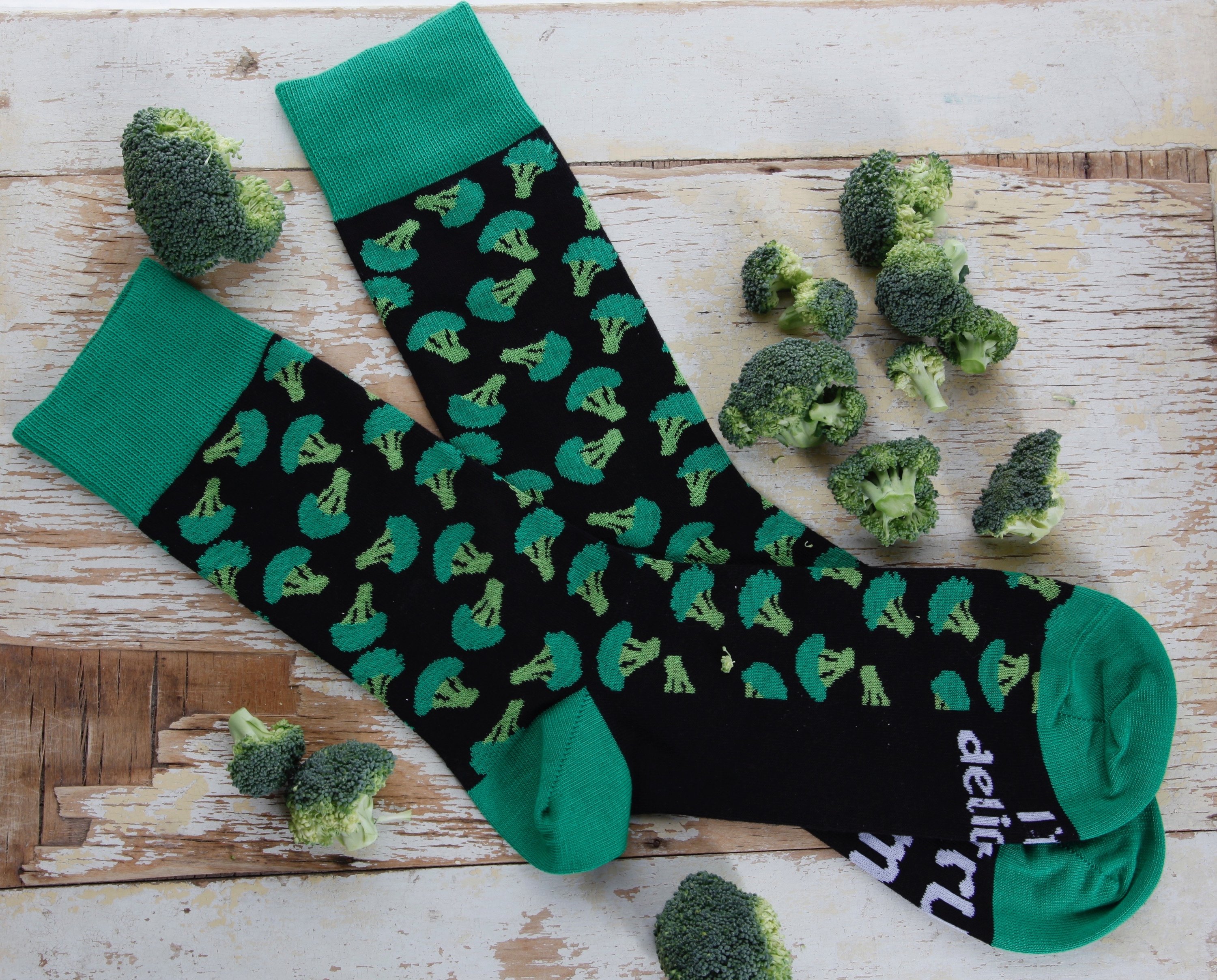 A pair of black cotton chef socks featuring a broccoli design and the phrase 'Trust me, I'm delicious' under the soles.