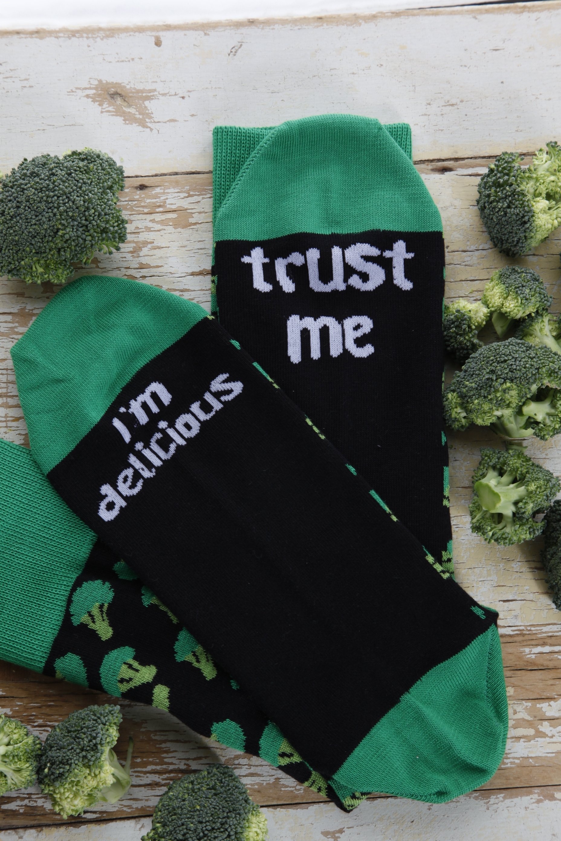 A pair of black cotton chef socks featuring a broccoli design and the phrase 'Trust me, I'm delicious' under the soles.