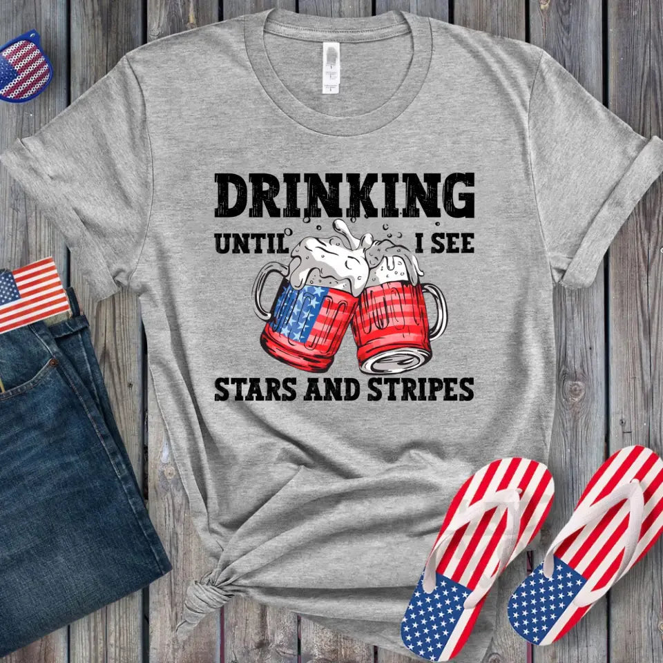 A stylish 4th of July tee featuring the phrase 'Drinking Until I See The Stars And Stripes' in a vibrant design, perfect for summer celebrations.