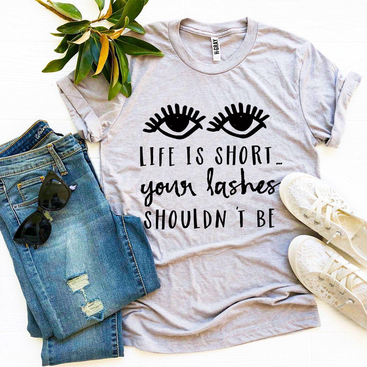 A stylish black t-shirt featuring the phrase 'Life Is Short Your Lashes Shouldn’t Be' in a modern font, made from soft ring spun cotton.