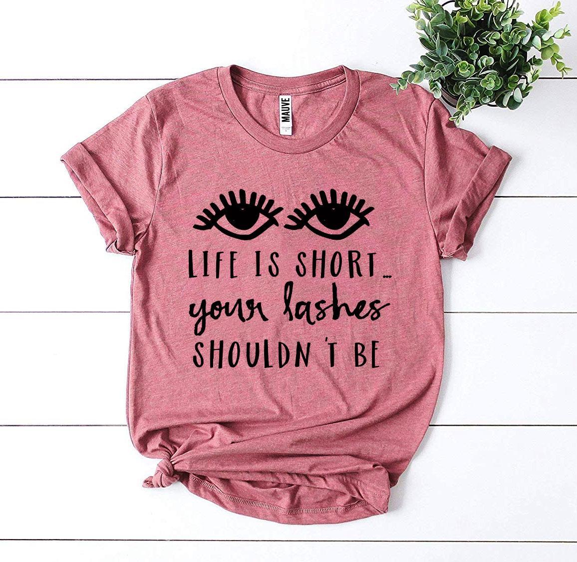 A stylish black t-shirt featuring the phrase 'Life Is Short Your Lashes Shouldn’t Be' in a modern font, made from soft ring spun cotton.