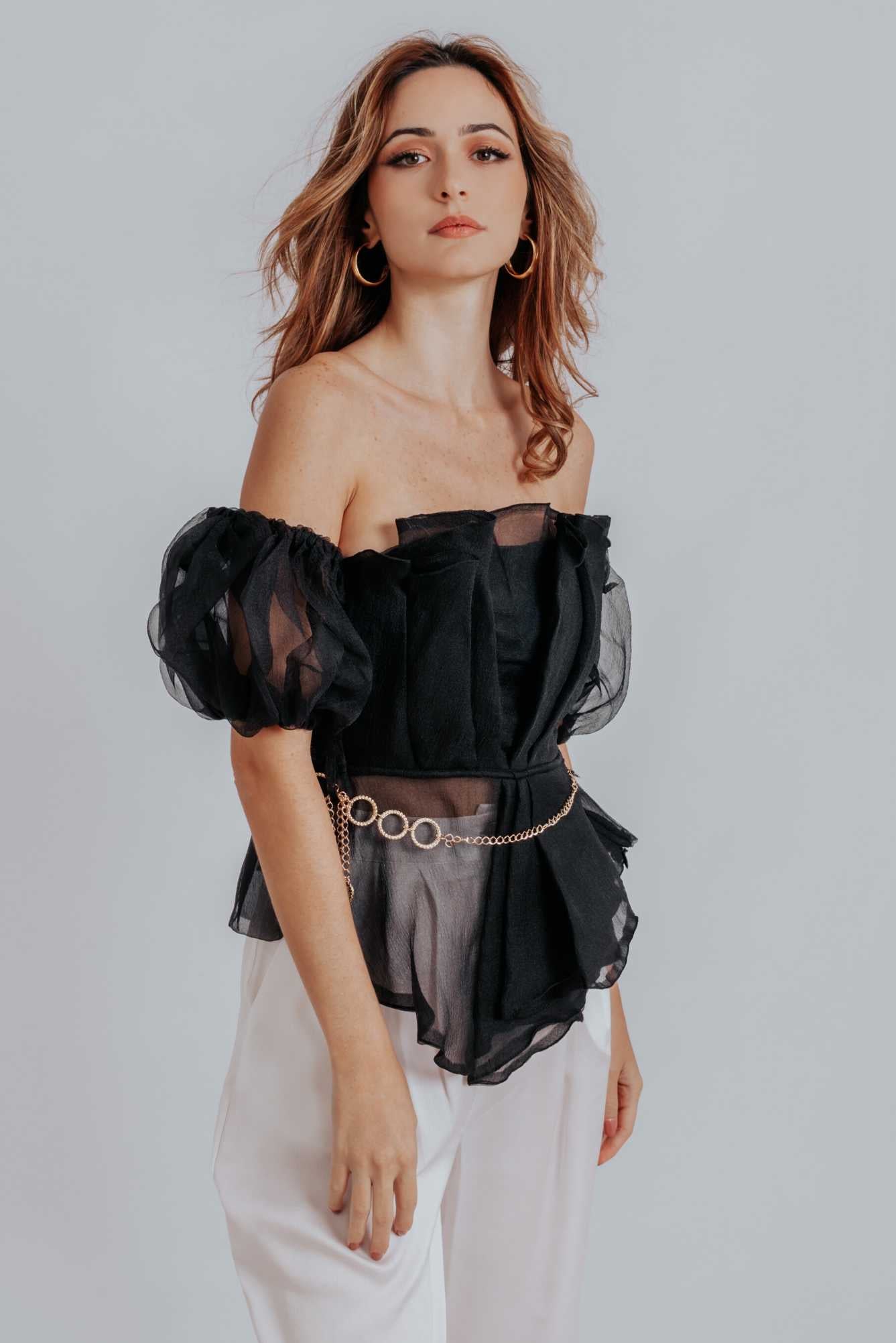 Raven Peplum Top featuring off-the-shoulder neckline and pleated bodice, styled with trousers and heels.