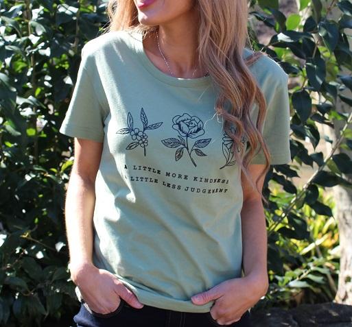 A Little More Kindness Tee in various colors, showcasing its casual graphic design and 100% cotton material.