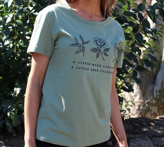 A Little More Kindness Tee in various colors, showcasing its casual graphic design and 100% cotton material.