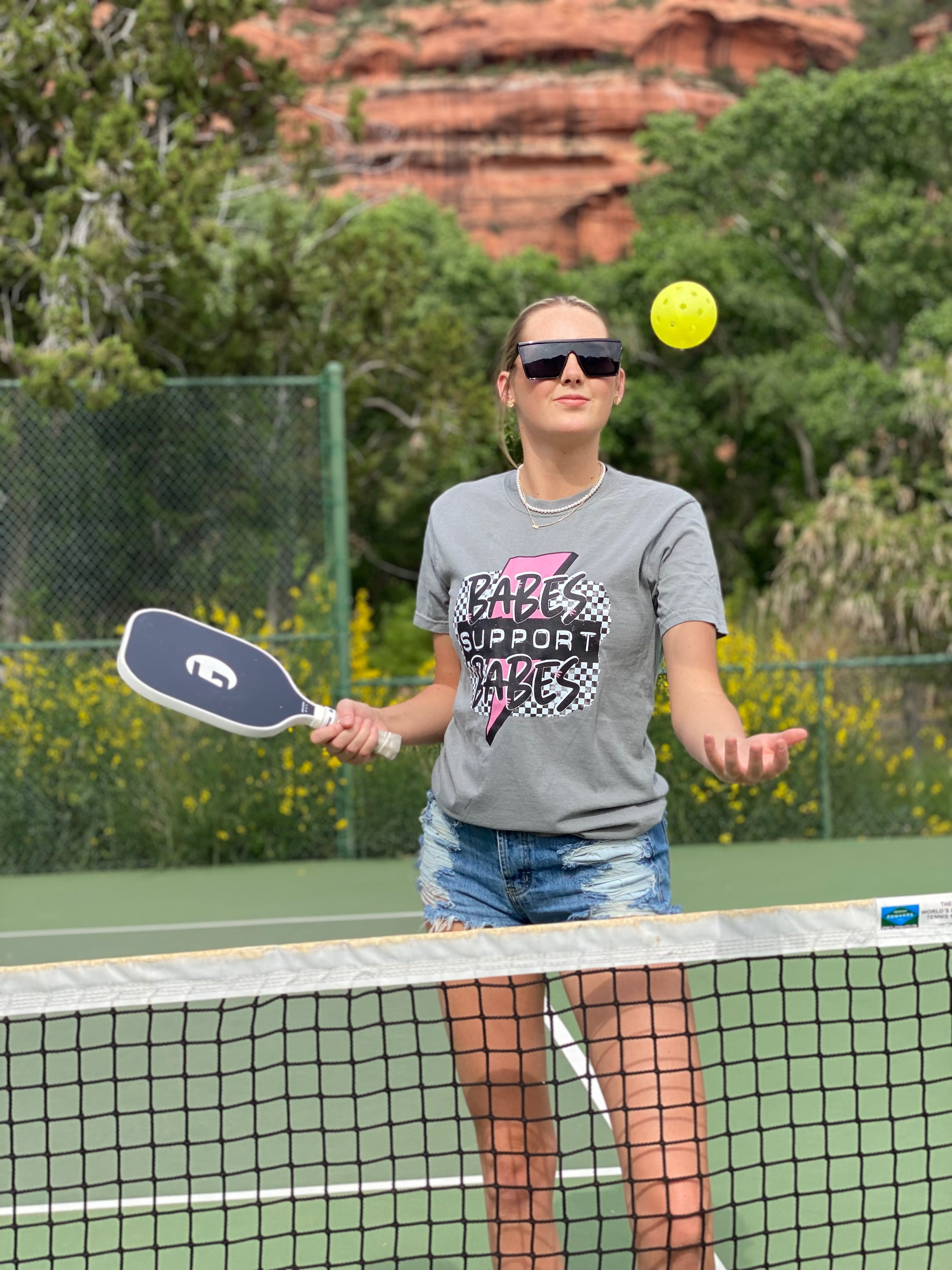 A gray graphic tee featuring the text 'Babes Support Babes' in a playful font, made from 100% cotton for comfort.