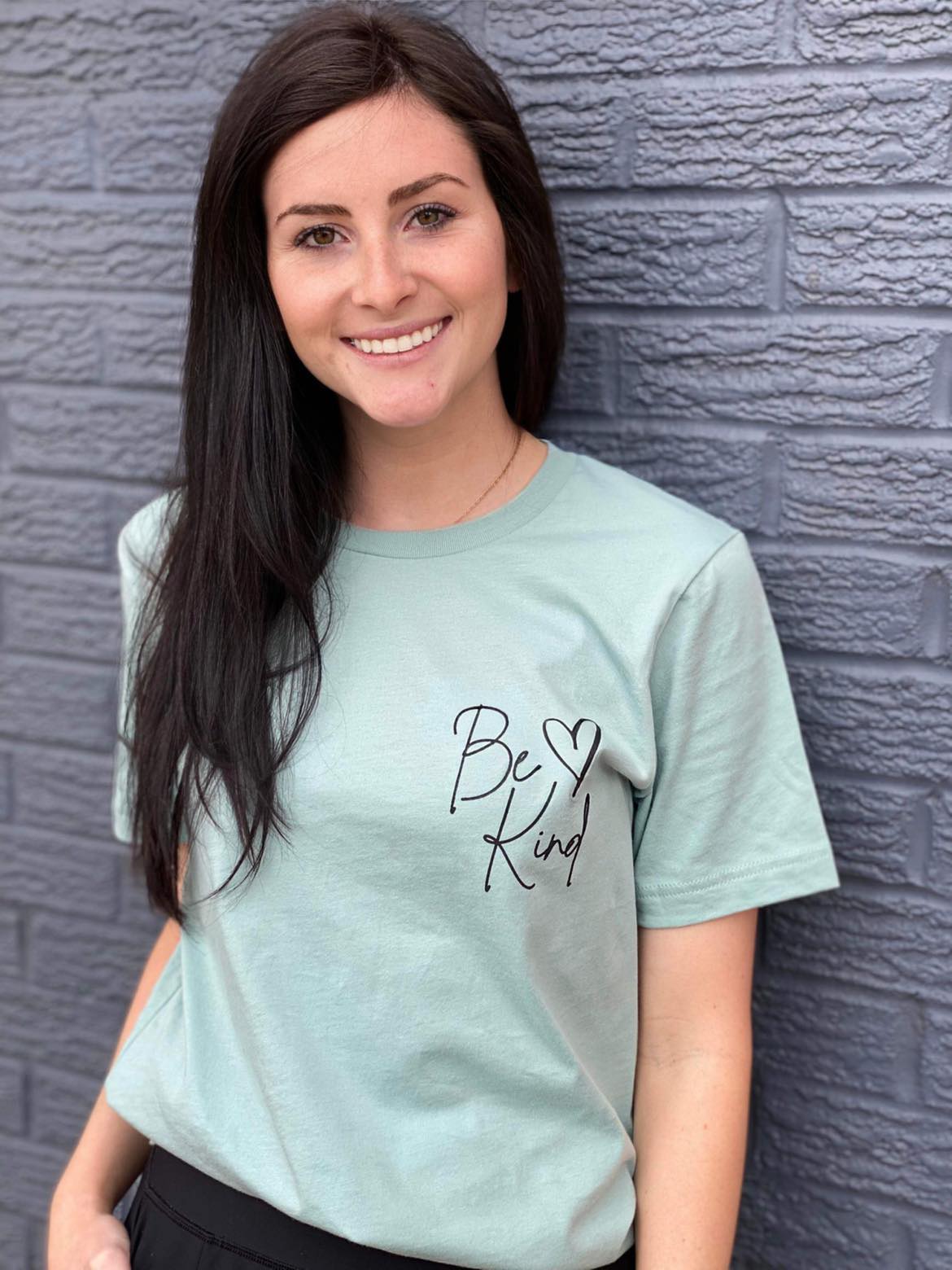 A soft Heather Dusty Blue tee with the message 'Be Kind Dear Person Behind Me' screen-printed in a stylish font, perfect for spreading kindness.