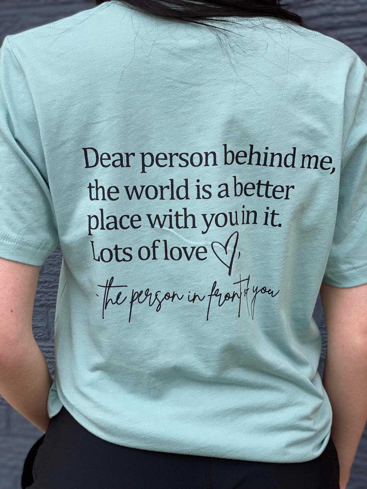 A soft Heather Dusty Blue tee with the message 'Be Kind Dear Person Behind Me' screen-printed in a stylish font, perfect for spreading kindness.