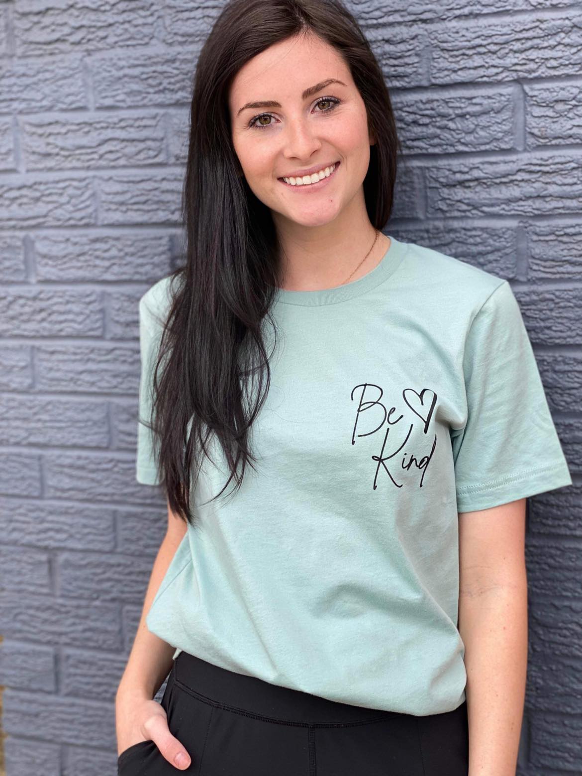 A soft Heather Dusty Blue tee with the message 'Be Kind Dear Person Behind Me' screen-printed in a stylish font, perfect for spreading kindness.