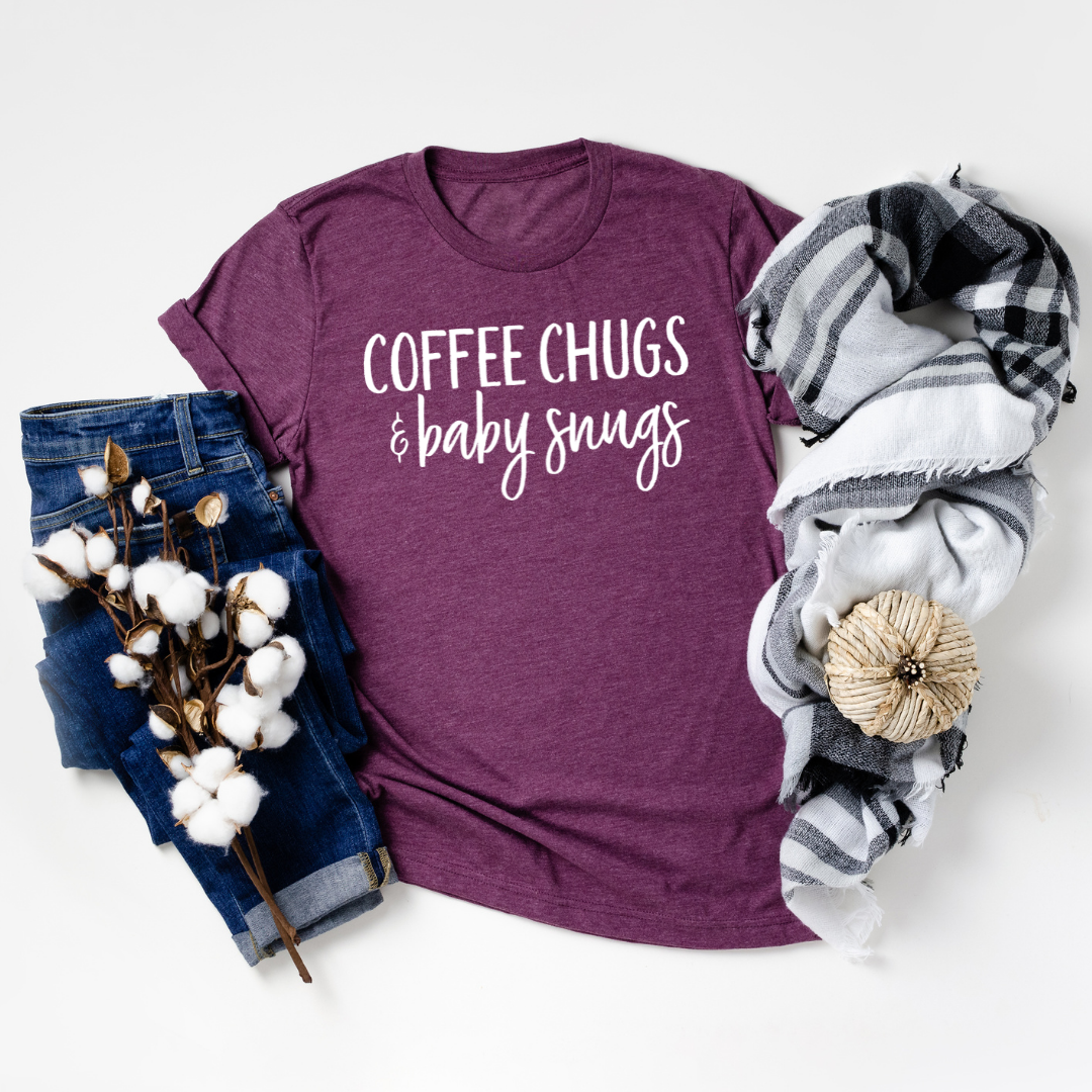 A soft, lightweight unisex T-shirt featuring the phrase 'Coffee Chugs & BABY Snugs' printed in white ink on a stylish background.