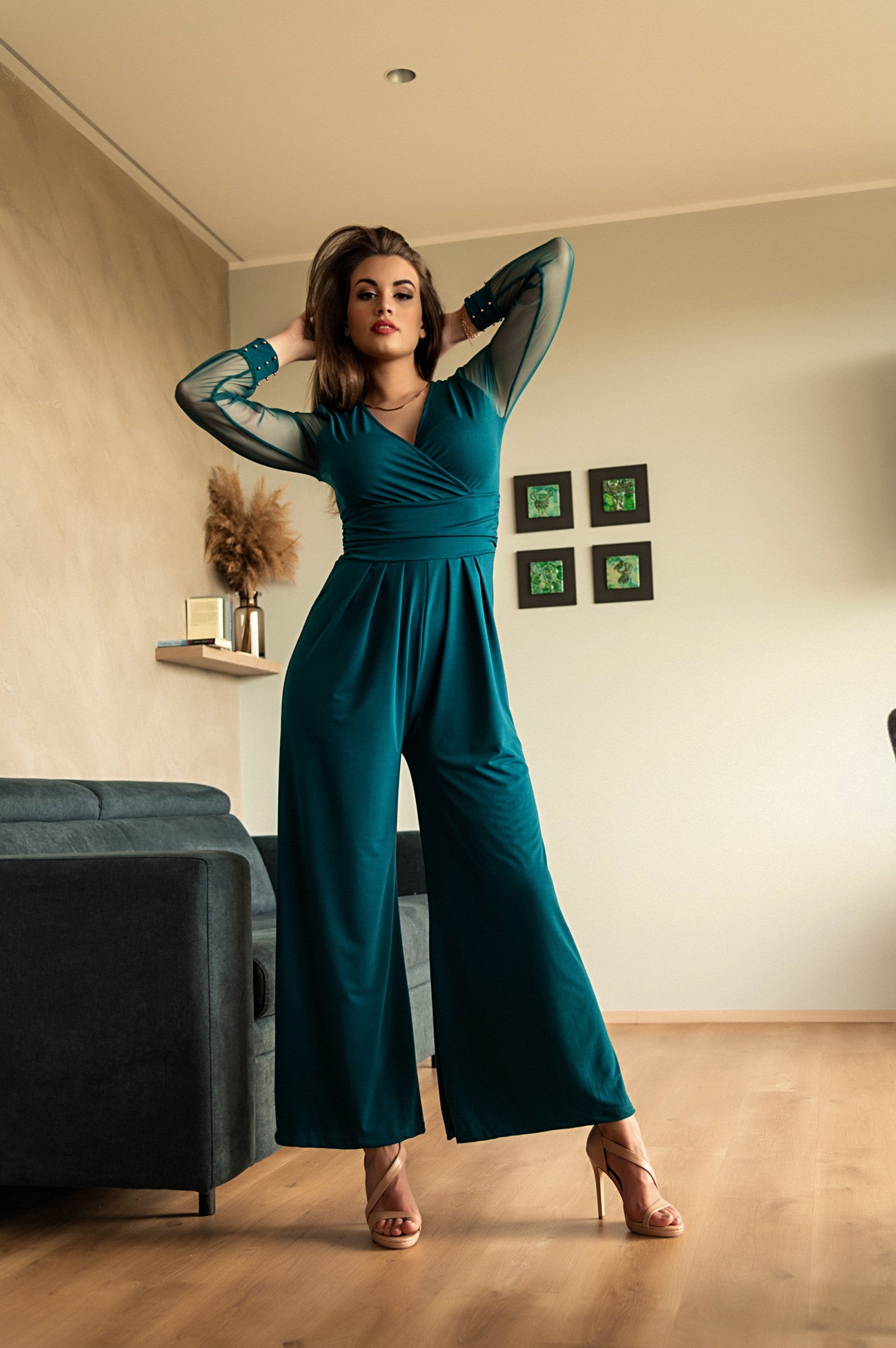 Elegant petrol jumpsuit with V neckline and long transparent sleeves, featuring gold rivets at the wrists.