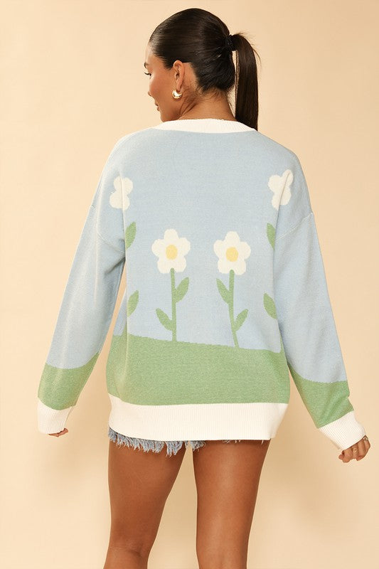 A stylish Flower Field Knit Cardigan featuring a vibrant flower field and sun intarsia design, button front, and V neckline.