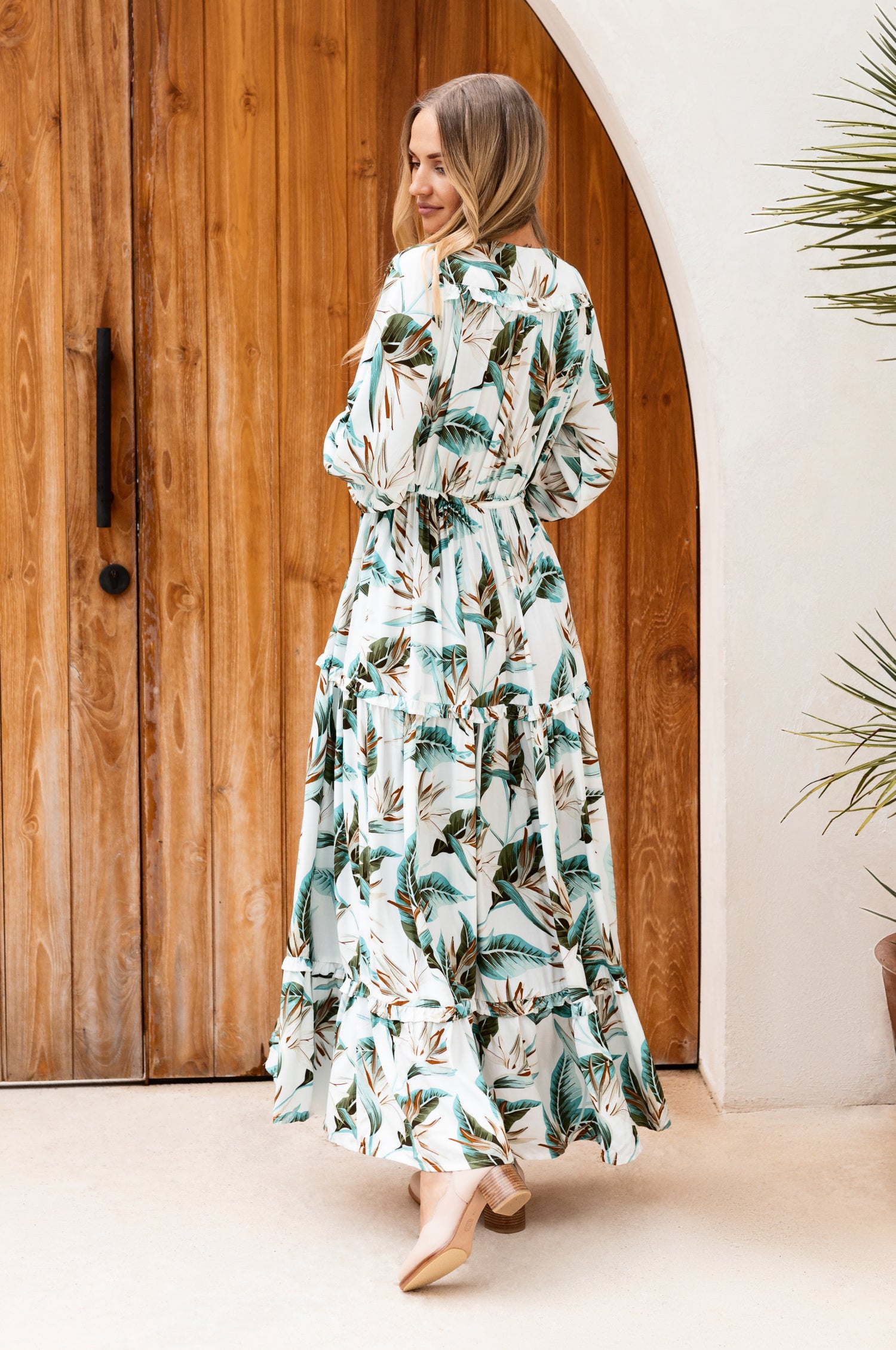 A beautiful woman wearing the Flower Fields Maxi Dress featuring a V-neckline, criss-cross tie, and ruffle details, surrounded by a vibrant floral backdrop.