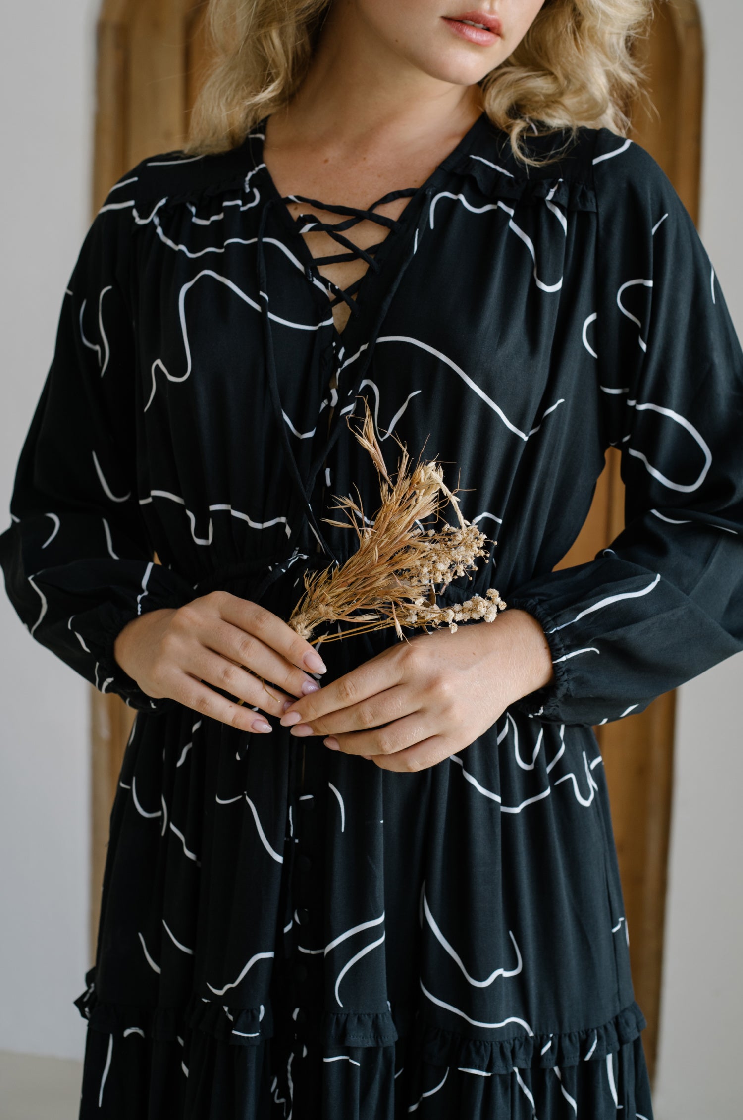 A beautiful woman wearing the Flower Fields Maxi Dress featuring a V-neckline, criss-cross tie, and ruffle details, surrounded by a vibrant floral backdrop.