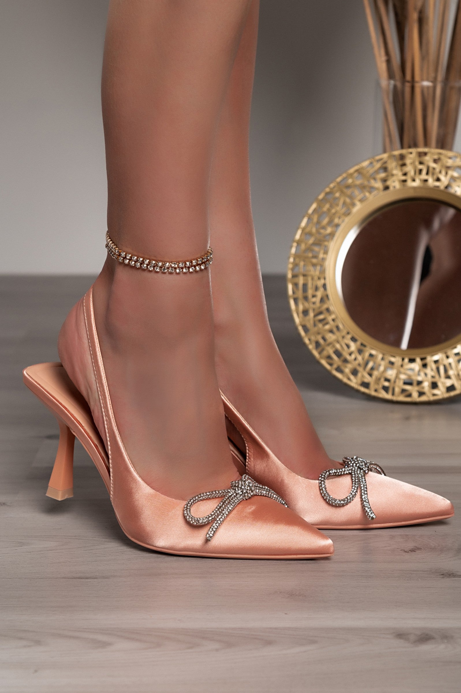 Beige high-heeled shoes with a decorative bow and pointed toe, featuring an elasticated strap for comfort.