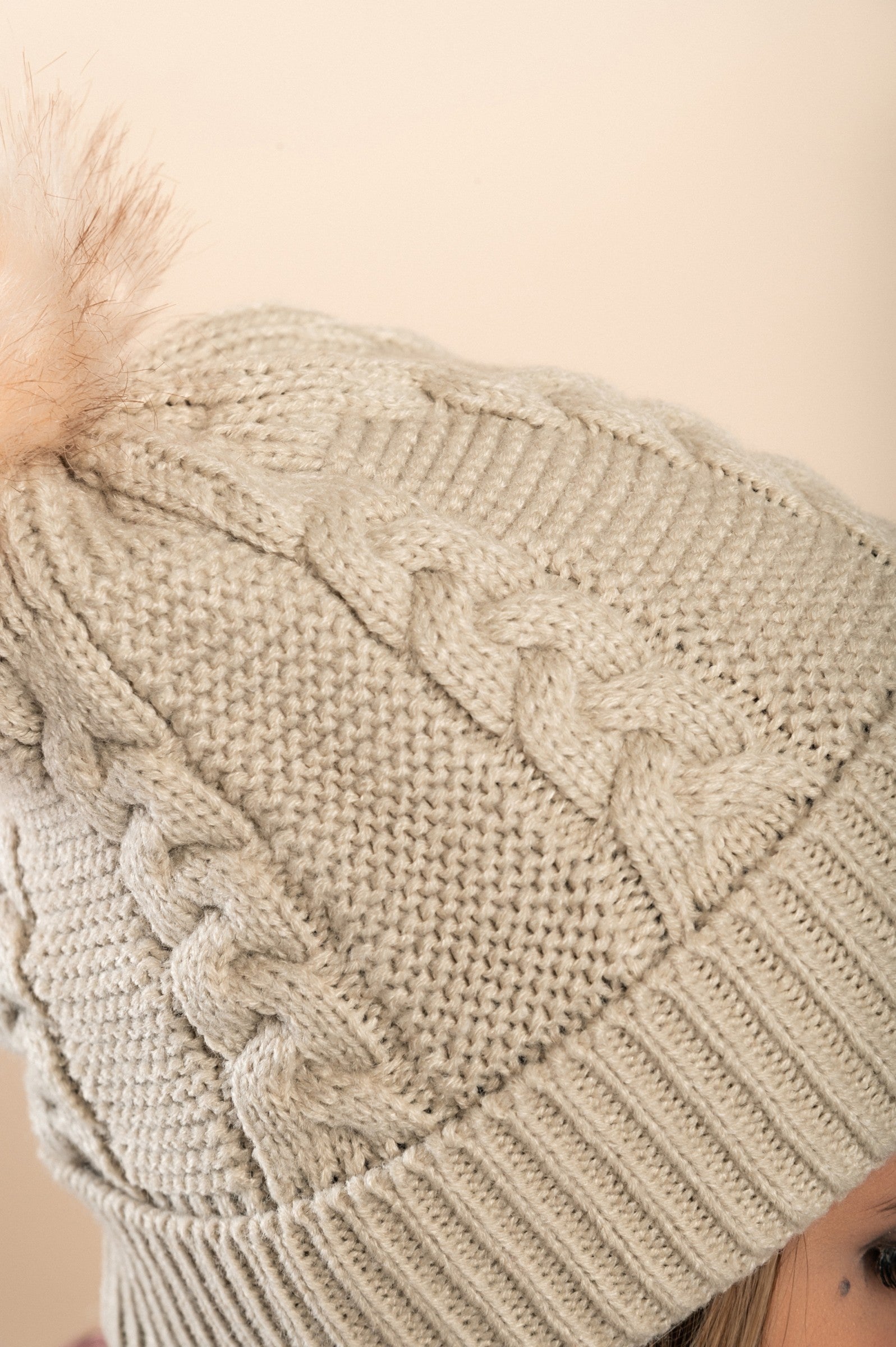 Beige knitted hat with a faux fur pompom on top, featuring a rolled edge and ribbed knit design.