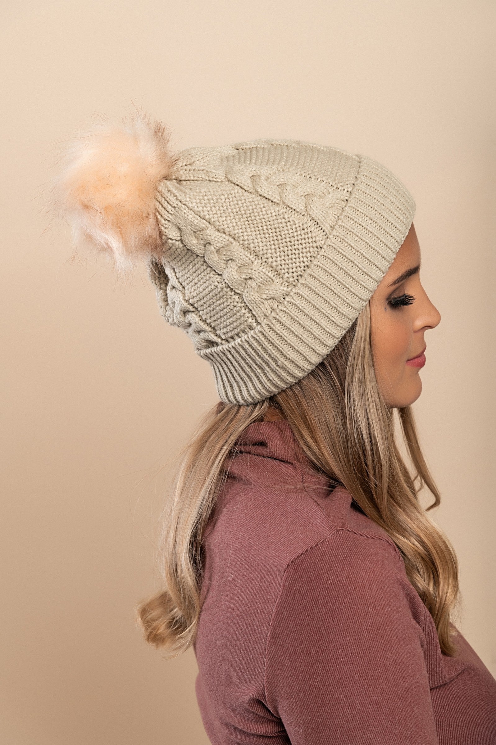 Beige knitted hat with a faux fur pompom on top, featuring a rolled edge and ribbed knit design.