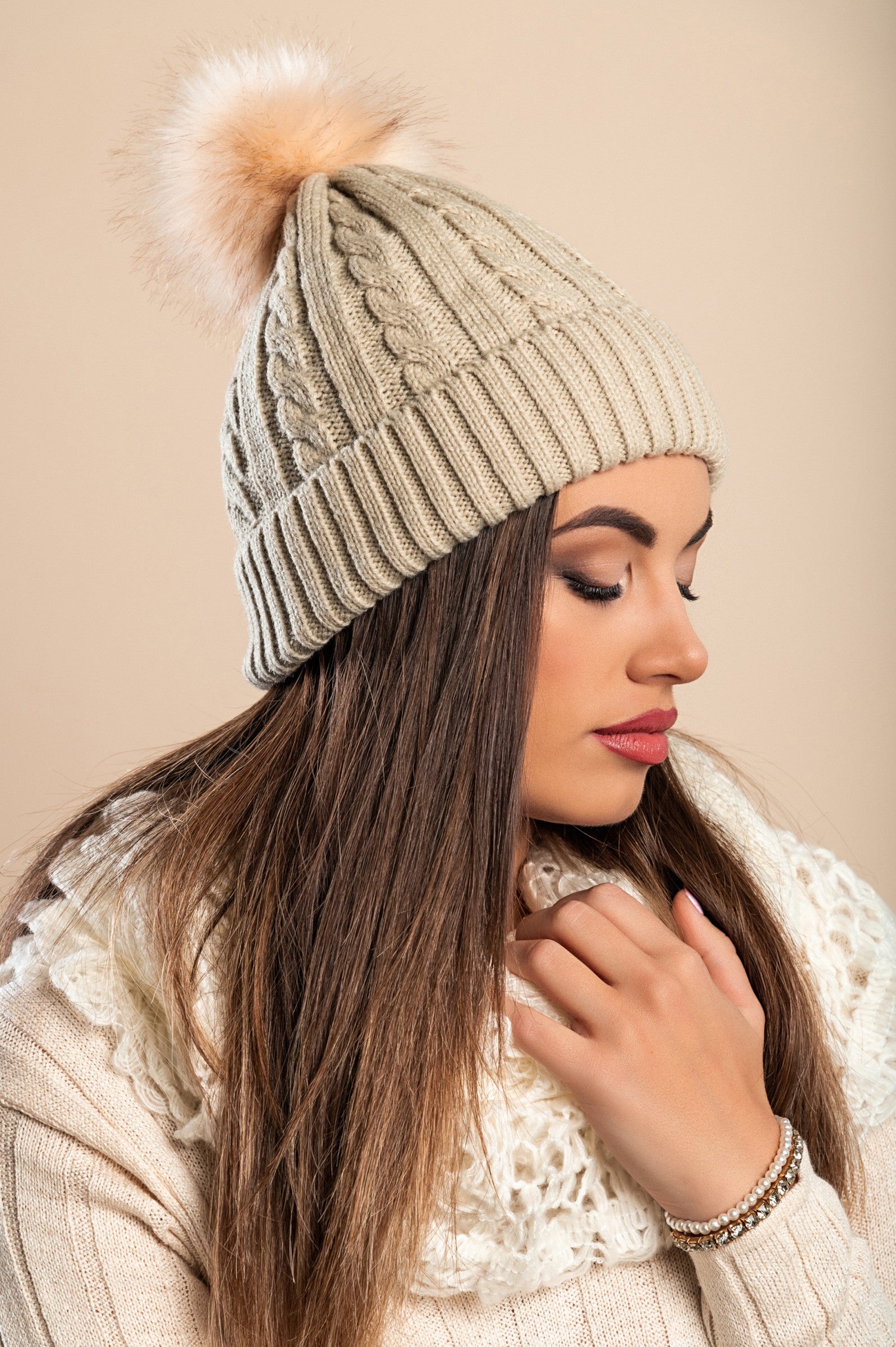 Beige knitted hat with a faux fur pompom on top, featuring a rolled edge and ribbed knit design.