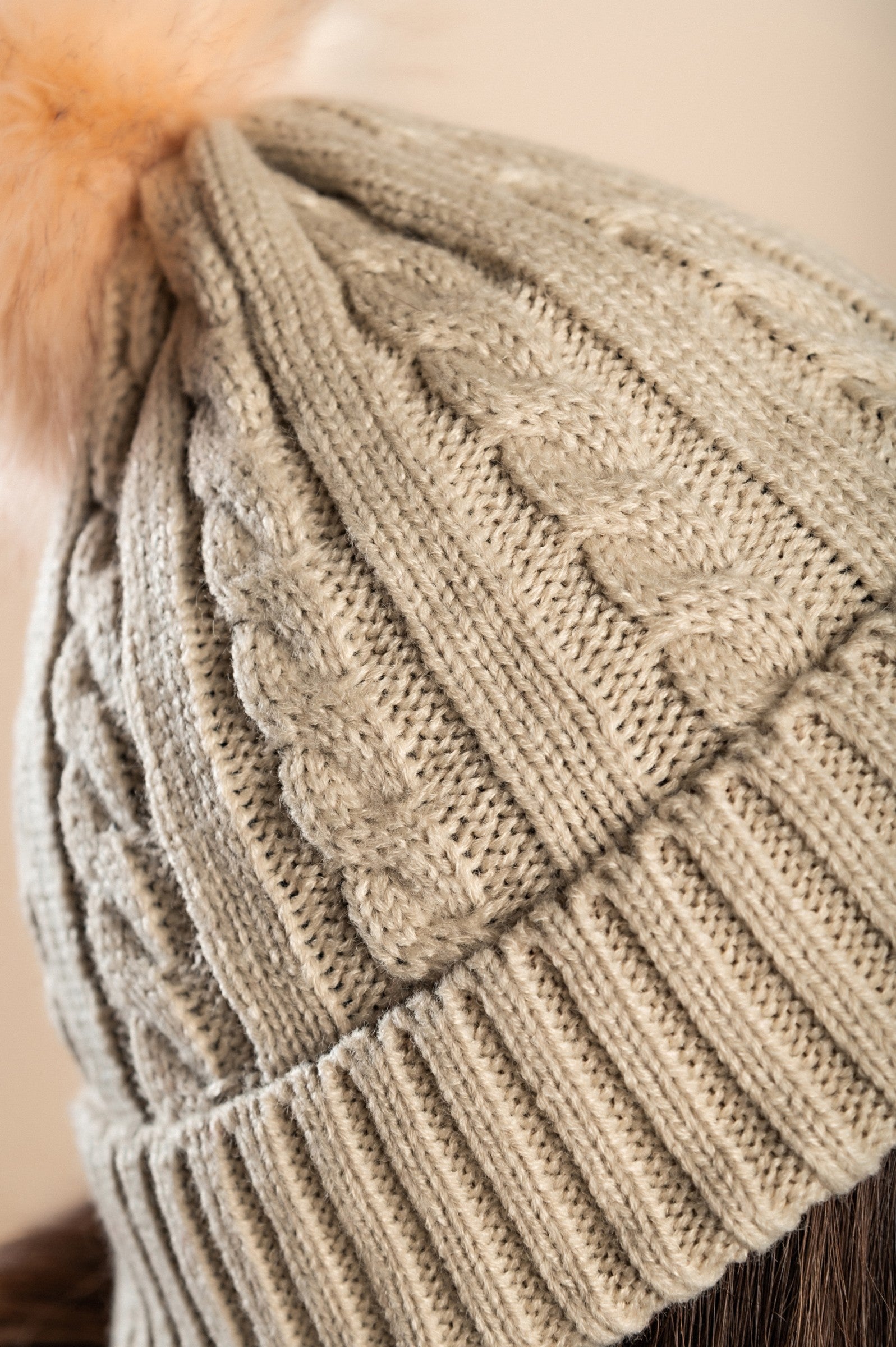 Beige knitted hat with a faux fur pompom on top, featuring a rolled edge and ribbed knit design.