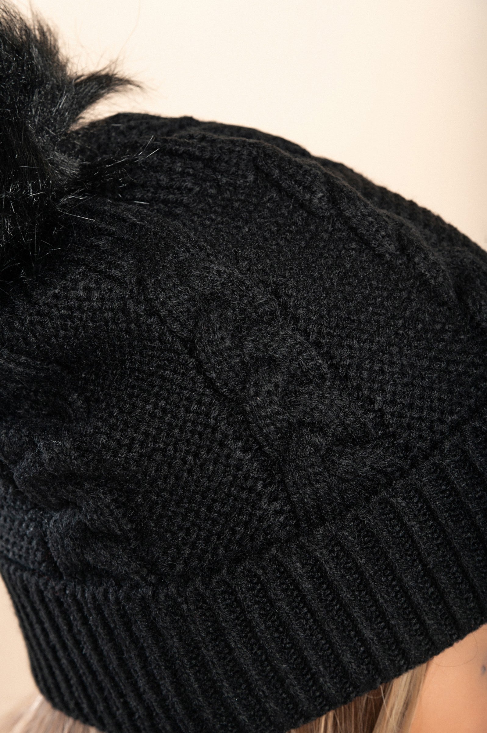 Black knitted hat with a faux fur pompom, featuring a rolled edge design, made from soft acrylic fabric.