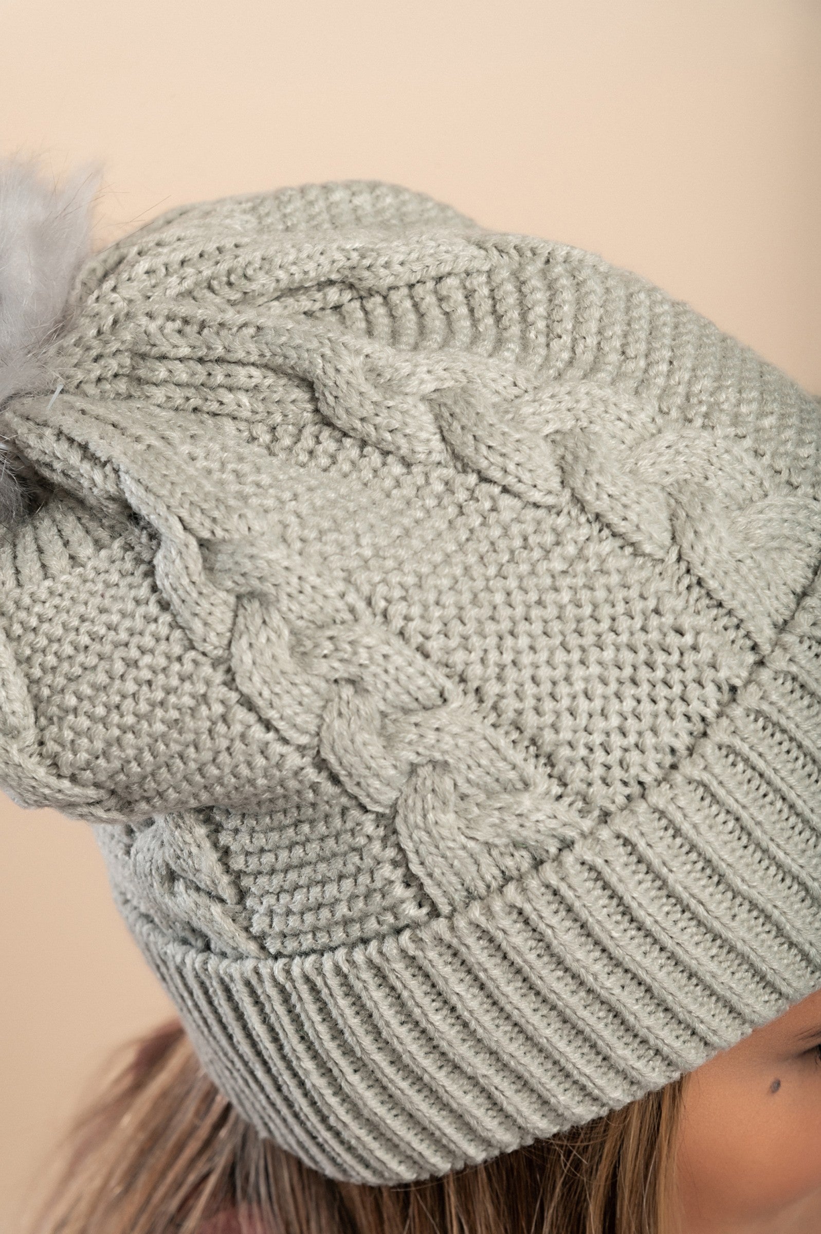 Light gray knitted hat with a faux fur pompom on top, featuring a rolled edge and ribbed knit design.