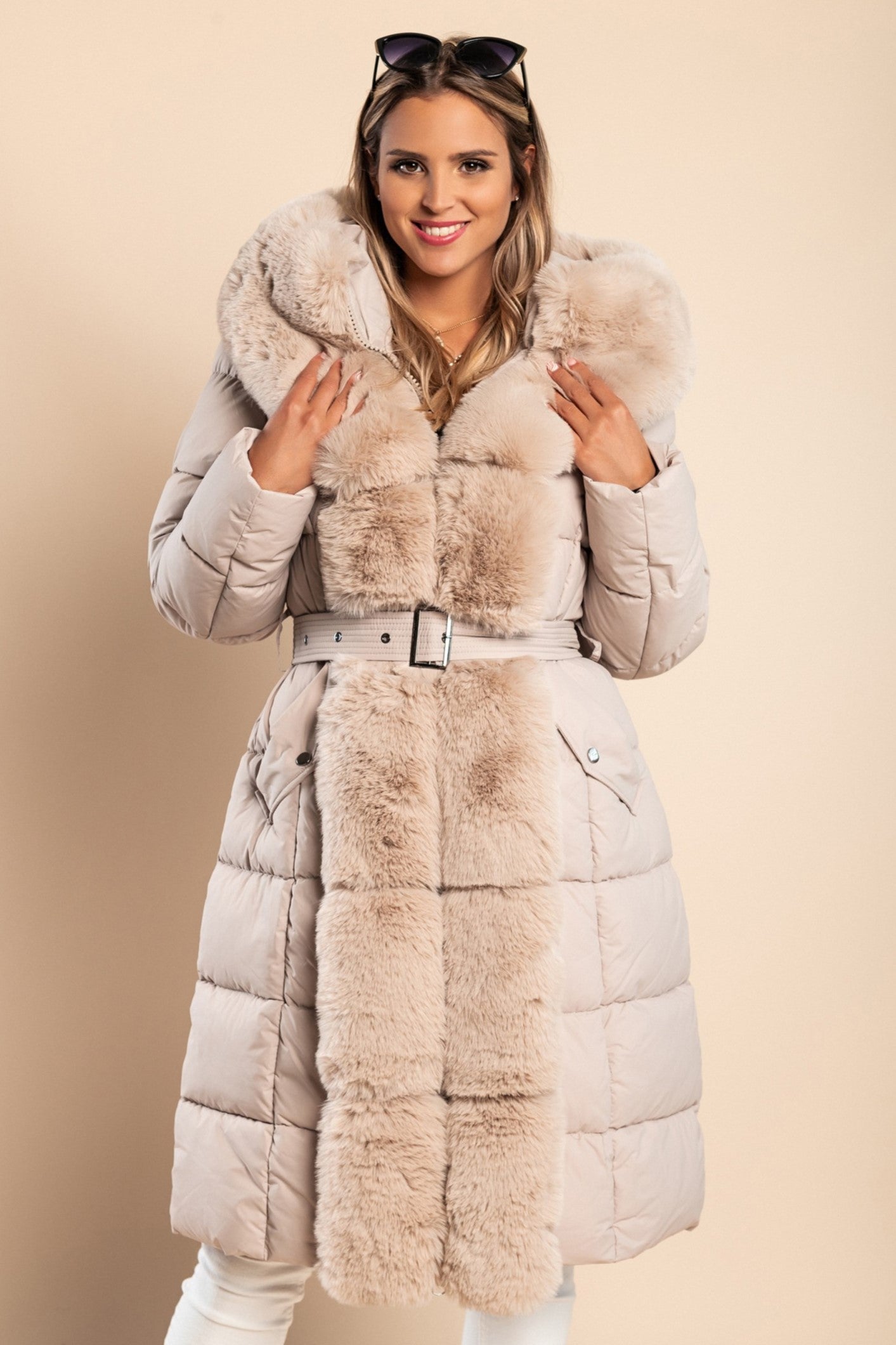 Beige padded winter jacket with removable faux fur hood and decorative belt, featuring side pockets and long sleeves.